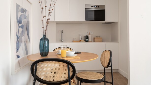 Een moderne keuken met witte kasten, een inbouwoven en een eethoek met een ronde tafel en rieten stoelen.