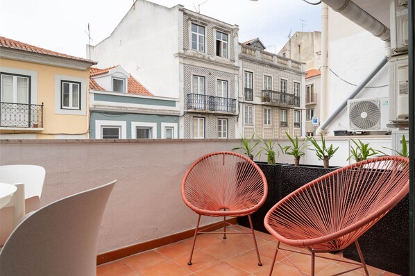 Un balcón con dos sillas de mimbre, una mesa blanca y plantas en macetas.