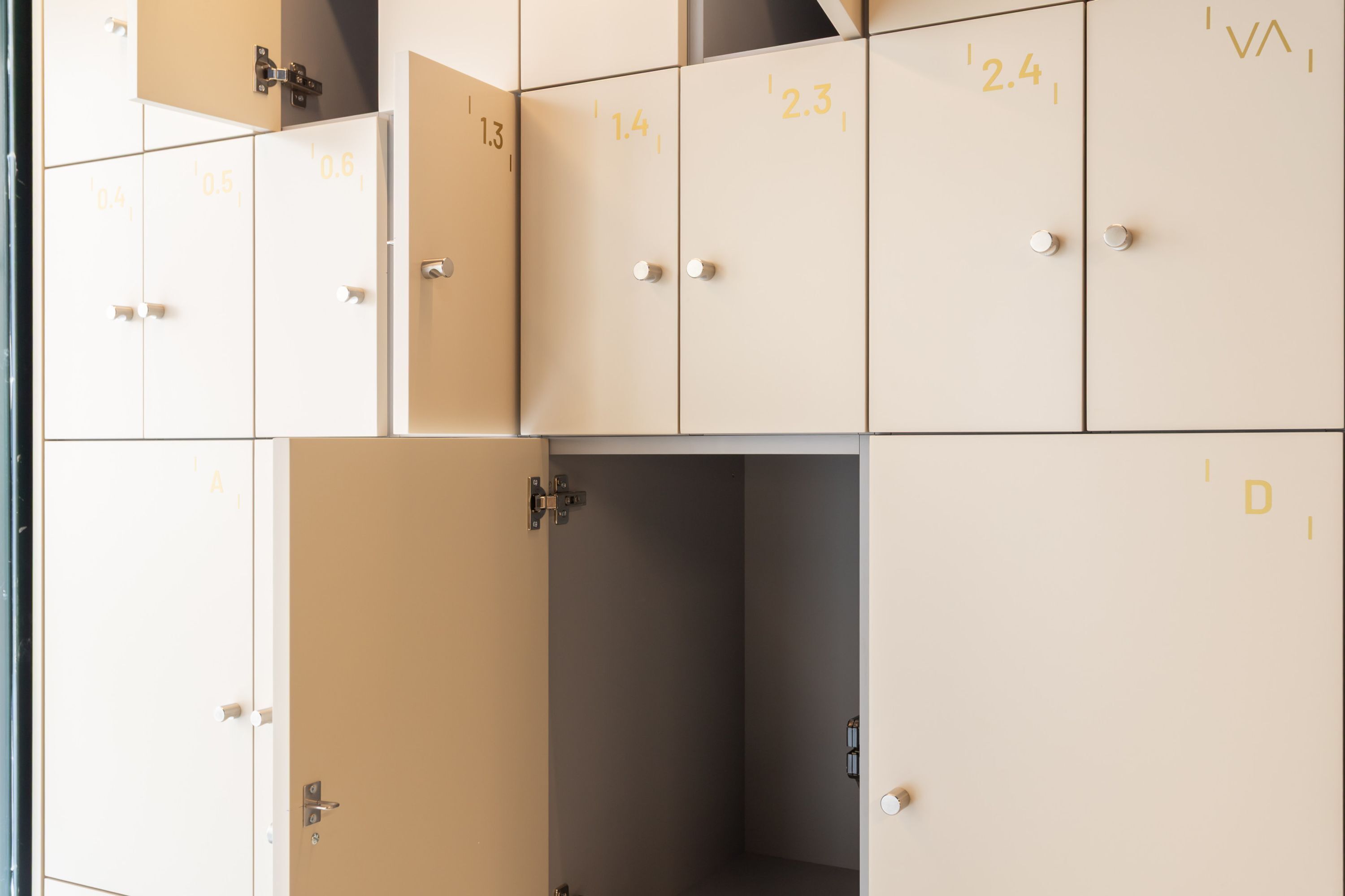 Una fila di armadietti beige con numeri da 0,4 a 2,4, alcuni sportelli aperti e una maniglia ben visibile.
