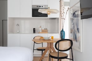 Une cuisine moderne avec des meubles blancs, une table à manger avec des chaises, une machine à café et un vase contenant des plantes séchées.
