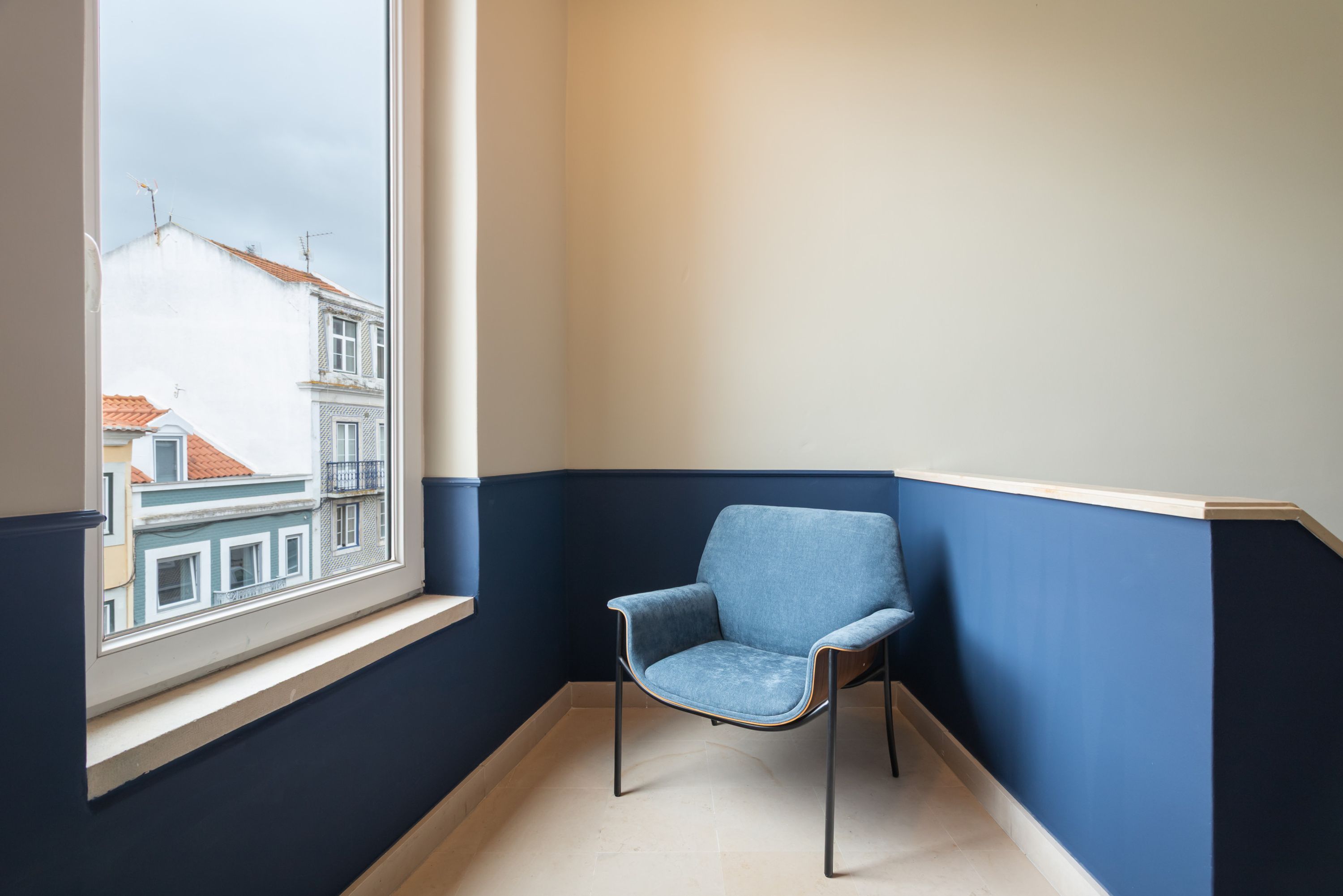 A corner room with a window, a blue chair, and a view of buildings outside.