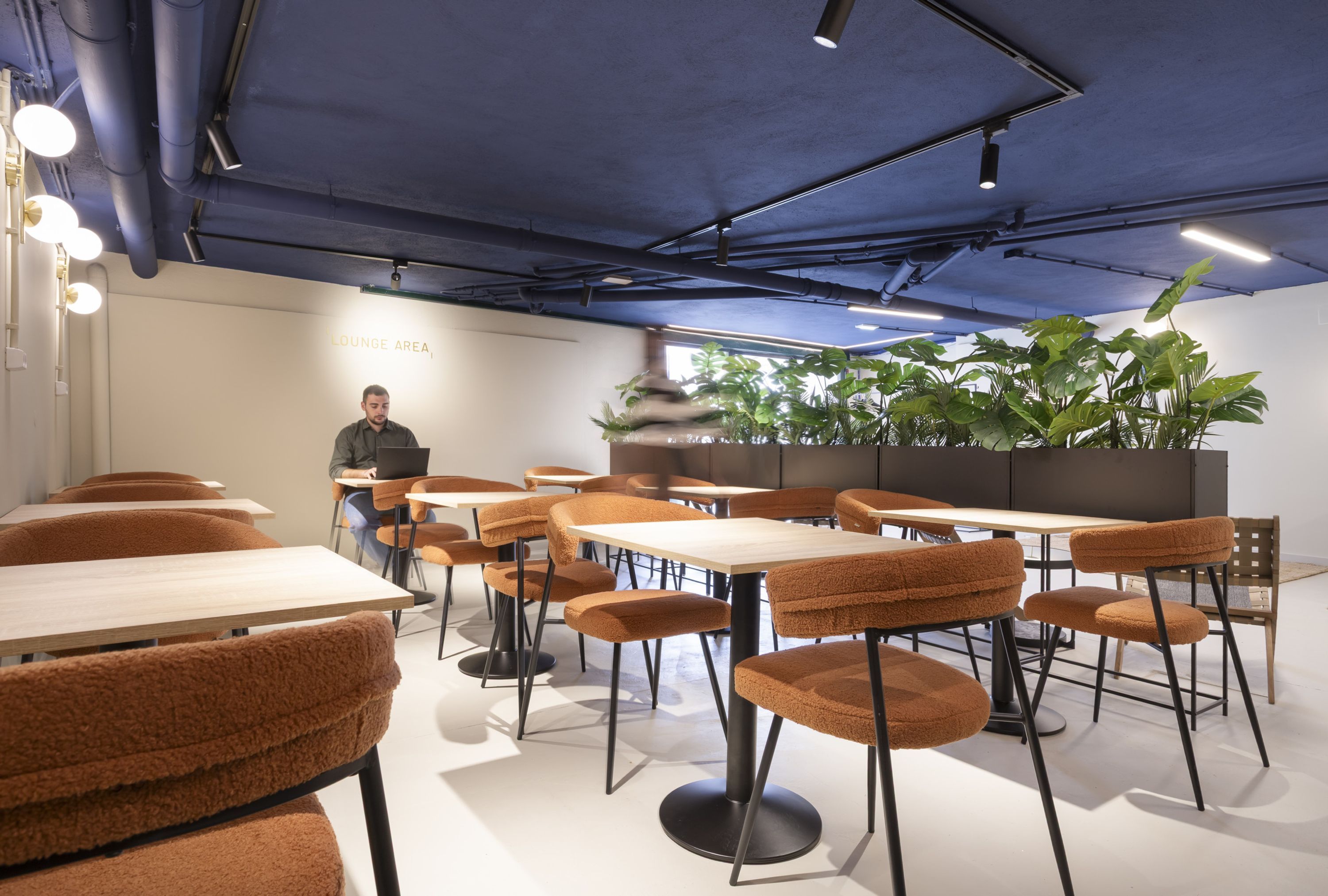 Een moderne loungeruimte met tafels en stoelen, een persoon zittend aan een tafel, en grote groene planten op de achtergrond.