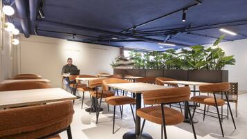 Een moderne loungeruimte met tafels en stoelen, een persoon zittend aan een tafel, en grote groene planten op de achtergrond.