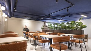 Un área de estar moderna con mesas y sillas, una persona sentada en una mesa y grandes plantas verdes al fondo.
