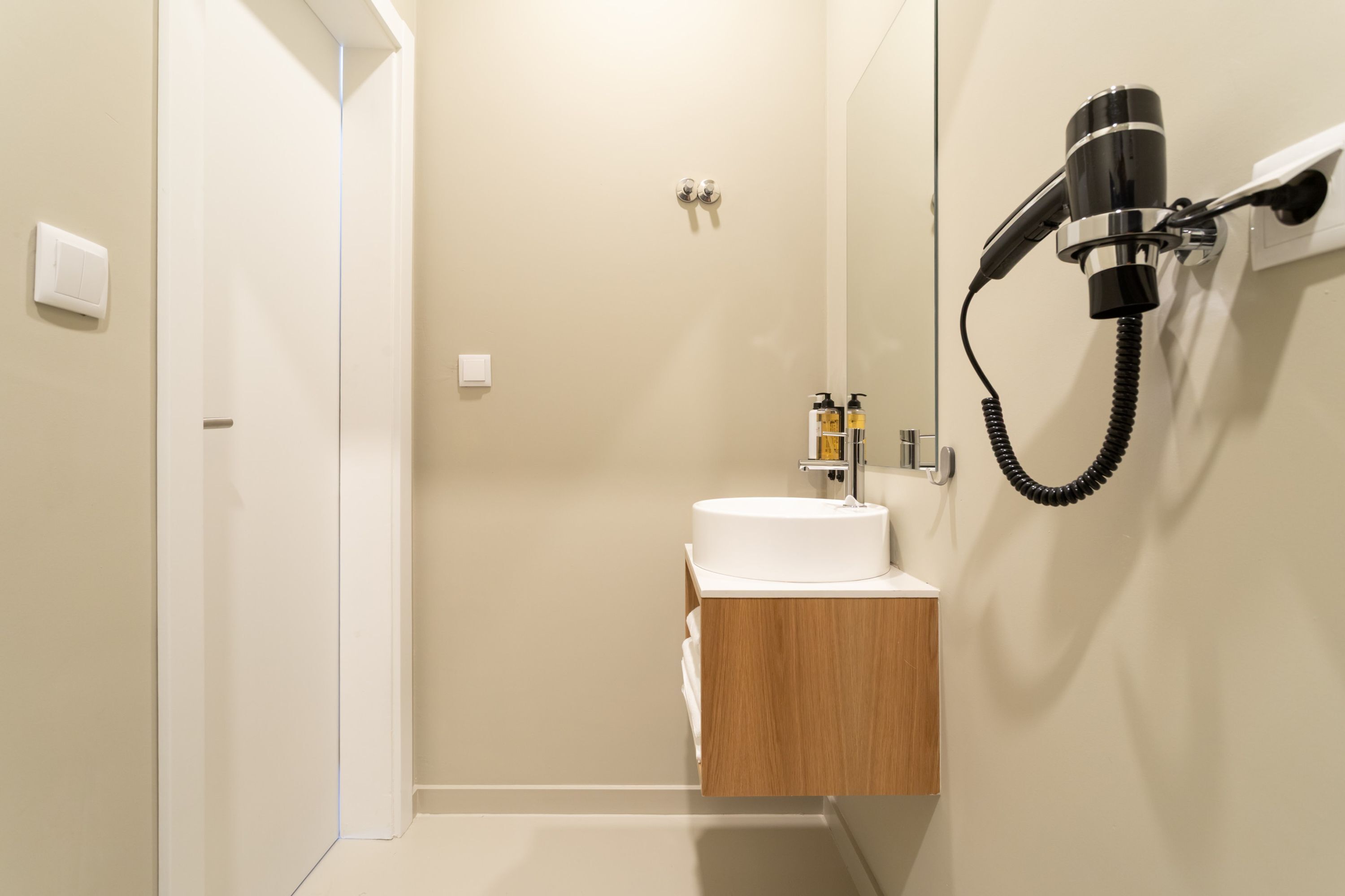 A bathroom with a white sink, a wooden vanity, a hairdryer mounted on the wall, and a door leading to another room.