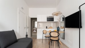 Un soggiorno moderno con divano, angolo da pranzo con sedie e tavolo, cucina con pensili e TV, e una porta che conduce in un'altra stanza.