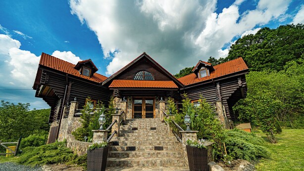 A large, rustic log cabin with a stone staircase leading to a wooden door, surrounded by lush greenery. - Rustic Cottage (Sibiu)