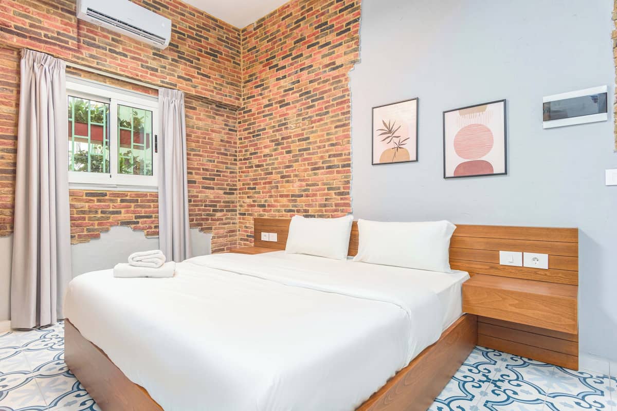 A hotel room with a brick wall, a large bed with white bedding, a window with curtains, and two framed artworks on the wall.