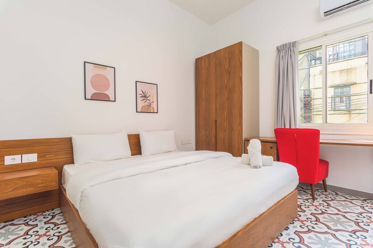 A hotel room with a bed, a wooden wardrobe, a desk with a red chair, and two framed pictures on the wall.