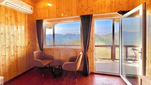 Habitación con paredes y pisos de madera, dos sillas, una mesita y ventanas amplias que ofrecen vista a la montaña.
