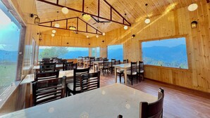 Un interior de madera espacioso con múltiples mesas y sillas, ventanas amplias que ofrecen vistas a la montaña y candelabros colgantes.