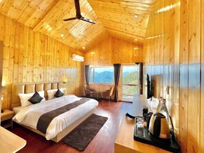A wooden cabin bedroom with a bed, a desk, a chair, and a view of the mountains. - Hilltop Haven Hotel And Resort (Kotdwara)
