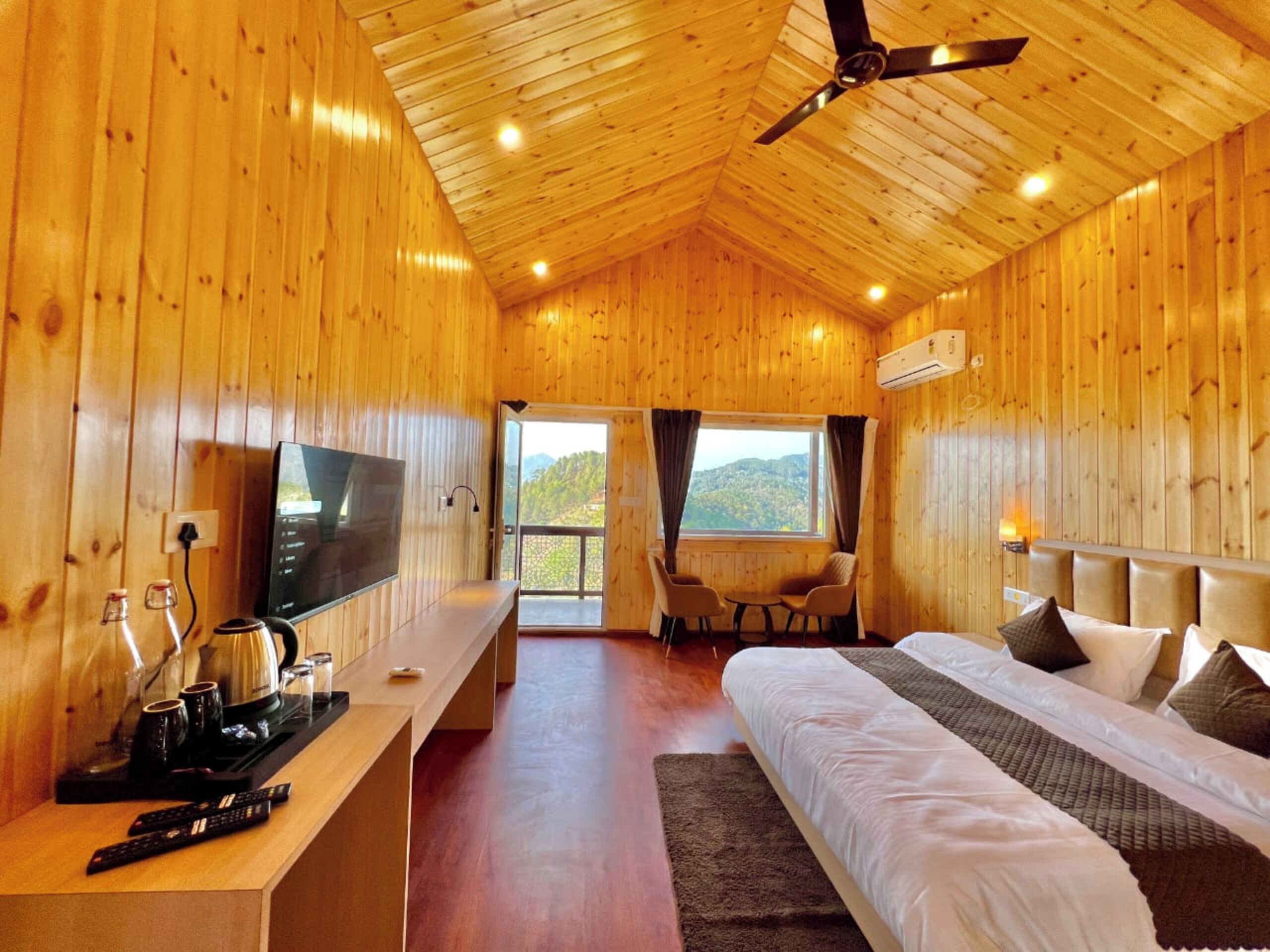 A wooden cabin room with a bed, a desk with a TV, and a balcony with chairs.