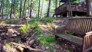 Una cabaña con terraza y banco cerca de un arroyo en un bosque.