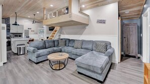 Una sala moderna con un sofá seccional gris, una mesa redonda de centro y un entrepiso con dormitorio y techo de madera.