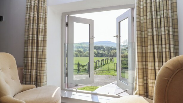 Une pièce avec deux fauteuils beiges, une grande porte vitrée donnant sur un balcon avec vue sur un paysage verdoyant et une clôture.
