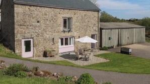 Une maison en pierre avec un toit en pente, un petit porche avec une table et des chaises, et un abri de jardin à l’arrière-plan.