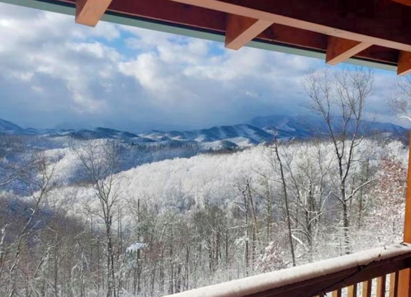 Blick vom Balkon mit Holzwertung auf eine verschneite Landschaft mit Bäumen und fernen Bergen.