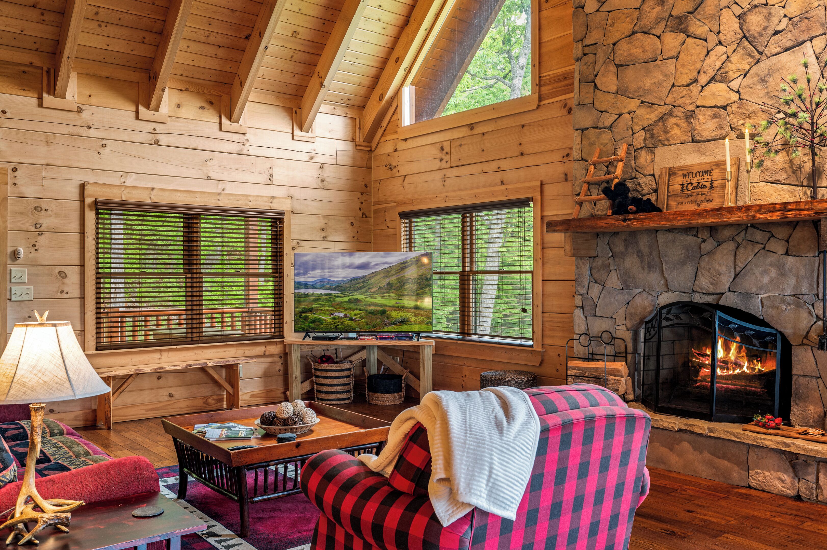 A cozy cabin interior with wooden walls, a stone fireplace, a television, and a large window with a scenic view.