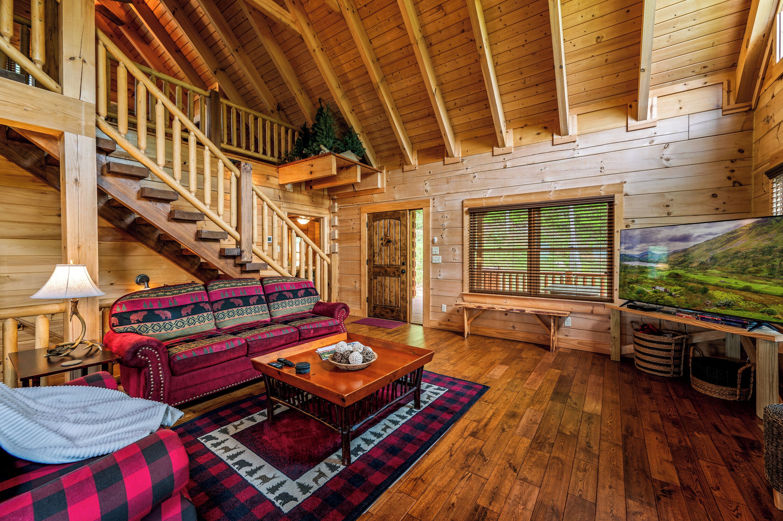 A log cabin interior with a wooden staircase, a sofa, a coffee table, and a large window with a view of a landscape.