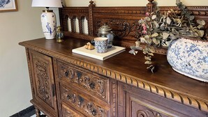 Uma cômoda de madeira com detalhes entalhados, um abajur, um vaso com flores e um prato decorativo.