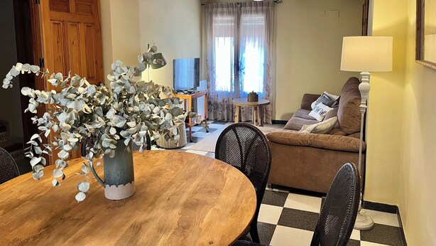 Uma sala de jantar com uma mesa de madeira, cadeiras e um vaso com folhas de eucalipto. Há um sofá e uma televisão ao fundo.