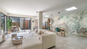 A spacious living area with a large sofa, dining table, and chairs. There is a kitchen area with cabinets and a mural on the wall.