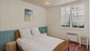 Ein Schlafzimmer mit einem hölzernen Bett, einem Nachttisch, einem kleinen Tisch, einem Heizkörper und einem Fenster mit Blick auf Grünflächen.
