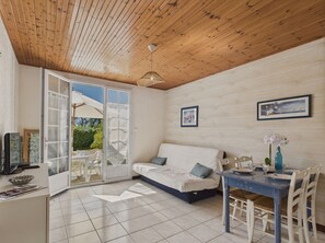 A cozy room with a wooden ceiling, a white sofa with blue cushions, a dining table with chairs, and a glass door leading outside. - 100 m from the Port – Apartment with Terrace for 4 (Talmont-Saint-Hilaire)