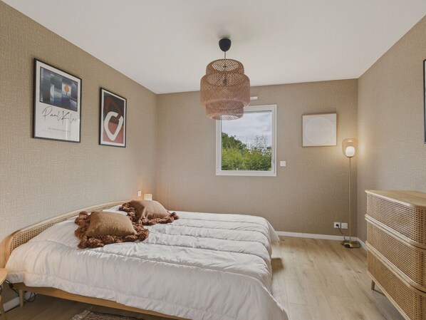 Une chambre avec deux lits, une fenêtre donnant sur un paysage verdoyant, un luminaire en fibres tressées et une table de chevet avec une lampe.