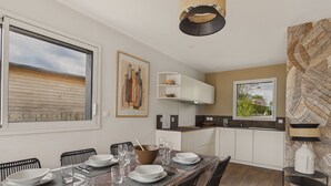 A modern kitchen with white cabinets, a dining area with a wooden table set for four, and a large window overlooking a wooden deck. - New house with terrace and garden for 6 (Fouesnant)