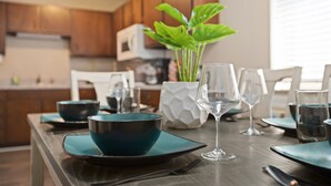 Mesa de jantar posta com tigelas verde-água, copos de vinho e pratos, com uma planta em vaso ao fundo.