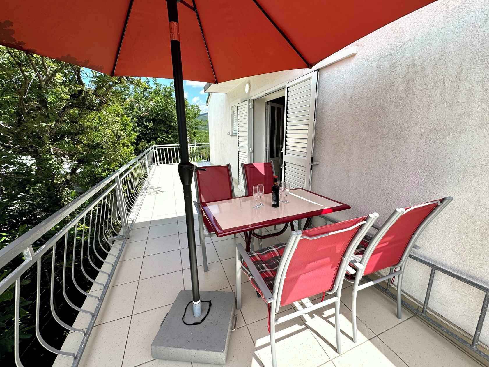 A balcony with a table and chairs, a red umbrella, and a view of trees and a building.