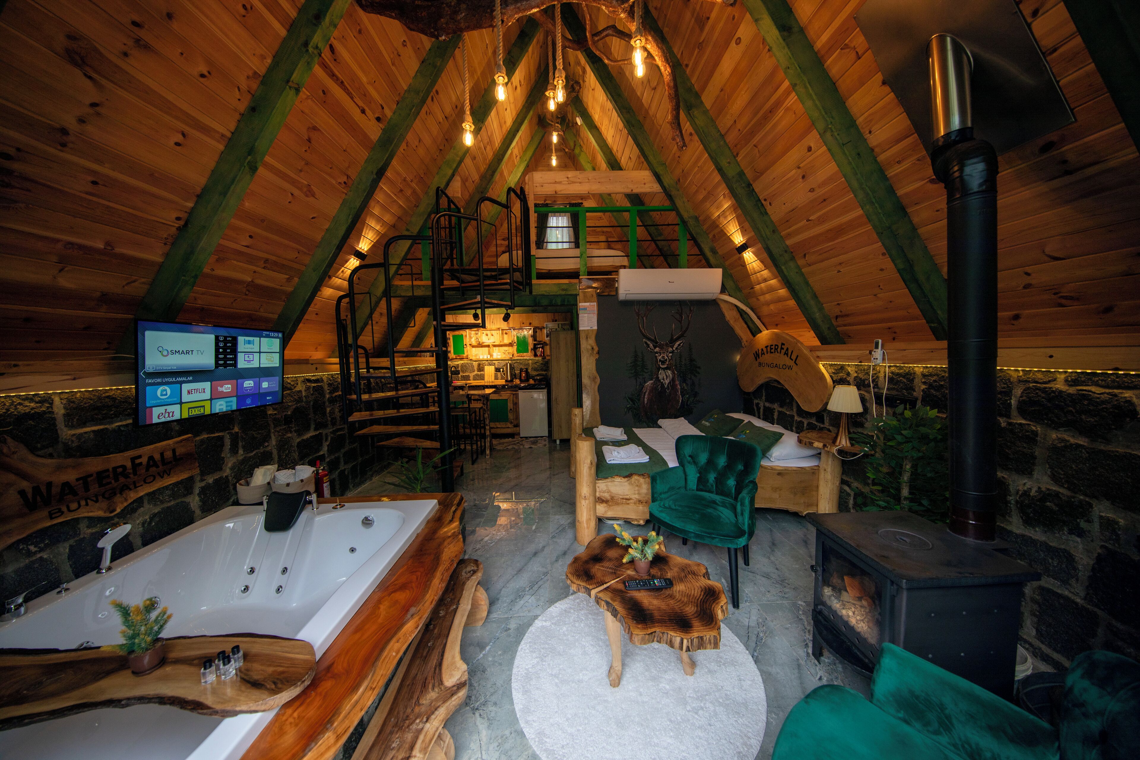 Een gezellige zolderkamer met een houten schuine dak, een groot bad, een groene stoel, een open haard en een televisie waarop een streamingdienst draait.