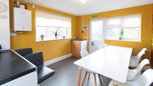 A yellow-walled room with a white table and chairs, a small kitchenette, and a window with a plant. - Thornbury Guest House Room 2 (Southampton)