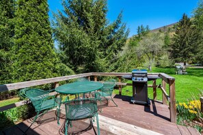 Eine Holzterrasse mit rundem Tisch und Stühlen, einem Grill und Blick auf eine grüne Rasenfläche mit Bäumen.