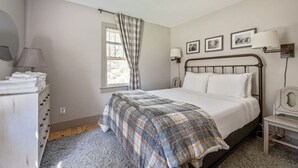 Een slaapkamer met een bed, een ladekast, een raam met gordijnen, wandlampen en ingelijste foto's.
