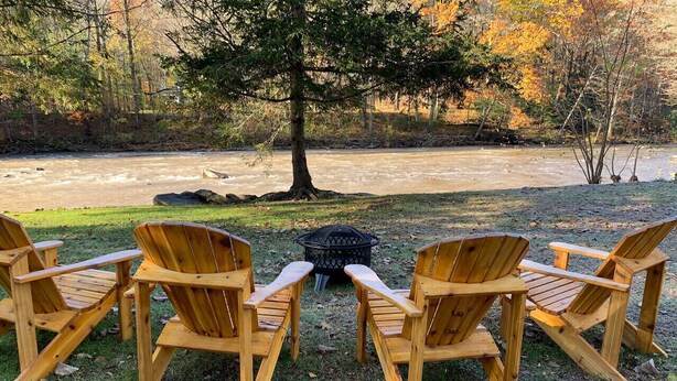 Four wooden Adirondack chairs facing a fire pit in a grassy area with a river and trees in the background. - "Rocky Bottom" Cabin • Creekside • Fire Pit • BBQ • Fishing (Phoenicia)