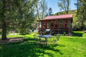 Een rustieke chalet met een rood dak, een houten terras en een eethoek met metalen stoelen en een tafel.