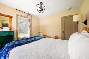 Een slaapkamer met een bed, een houten stoel, een bureau met handdoeken, een raam met gordijnen en een deur.