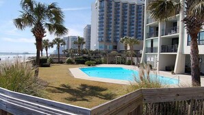 Ein Schwimmbecken, umgeben von Palmen und einer Holzterrasse, mit Blick auf den Strand und hohe Gebäude im Hintergrund.
