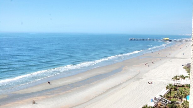 Ein Strand mit einem Steg, der sich ins Meer erstreckt, feiner Sand und einige Personen.