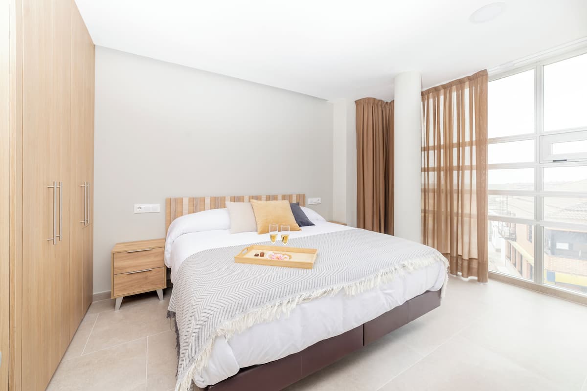 A modern bedroom with a large bed, wooden wardrobe, bedside tables, and a large window with curtains.