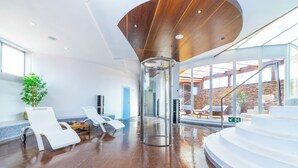 A modern indoor space with a wooden ceiling, a glass elevator, white lounge chairs, and a staircase leading to an outdoor area.