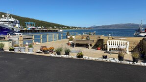 A waterfront dock with a blue ship, wooden benches, potted plants, and a clear view of the water and distant hills.