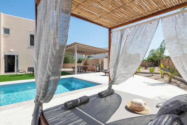 A poolside area with a pergola, a lounge chair, a sunbed, and a hat on the ground.