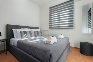 A modern bedroom with a large bed, a bedside table, a round ottoman, and a window with blinds.