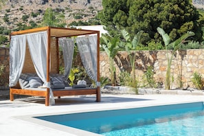 A wooden four-poster bed with a canopy, a swimming pool, and a stone wall with vegetation.