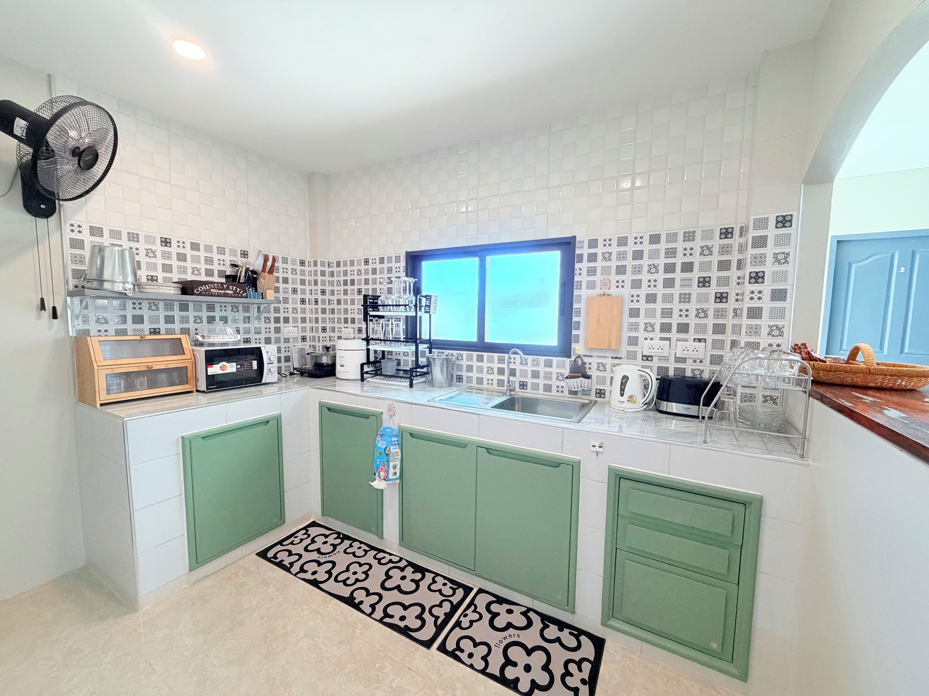 A kitchen with white tiled backsplash, green cabinets, a window, and a fan.