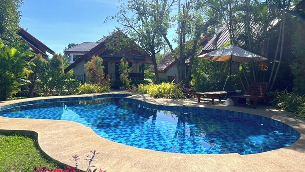 A swimming pool with a tiled edge, surrounded by tropical plants and a house with a tiled roof. - Siri Lagoona Resort & Pool Villa (Bang Lamung)